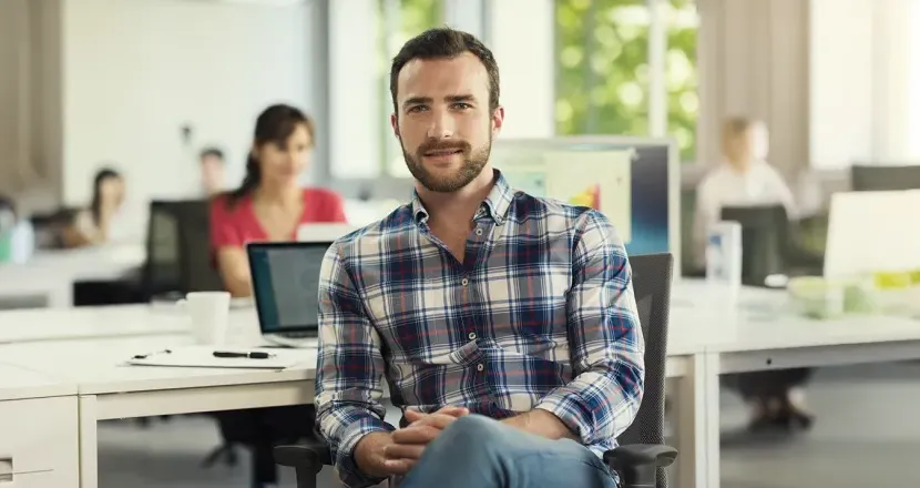 A Herzing University interdisciplinary studies student in a casual plaid shirt sits confidently in a modern, collaborative office environment, ready for a versatile career path.